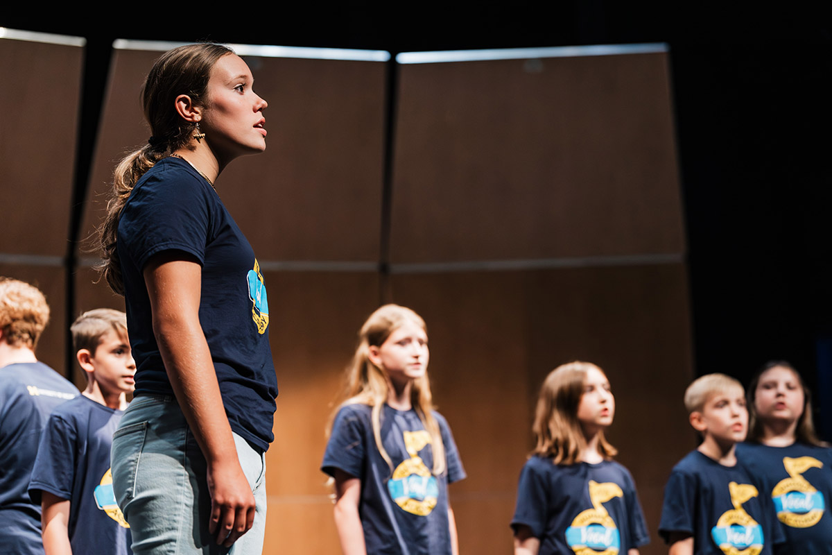 A young female student singing onstage with a group of vocalists behind her during the Summer Vocal Academy of Music.