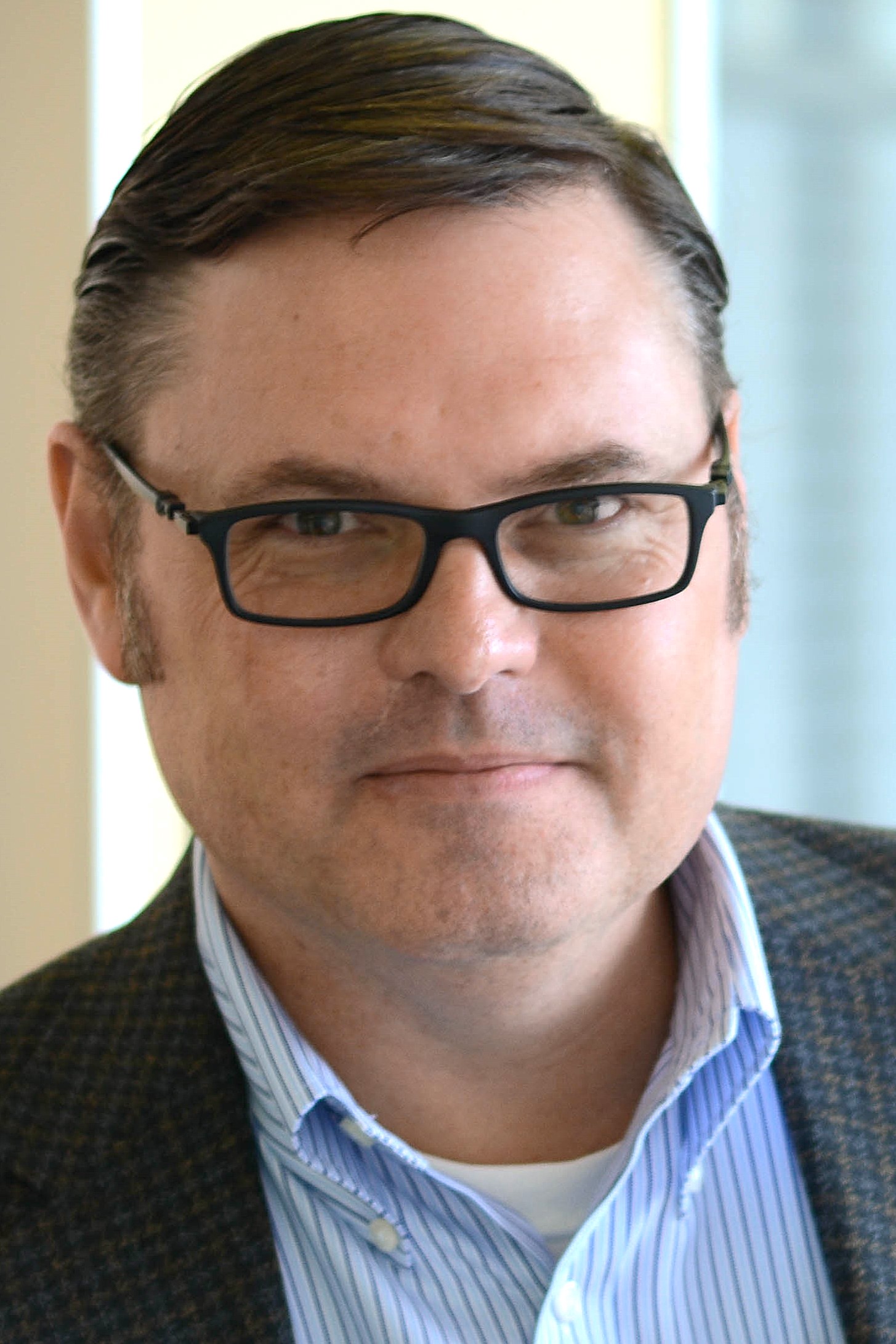 A man with short, neatly combed brown hair is wearing glasses, a dark checked blazer, and a collared blue-and-white, striped shirt.