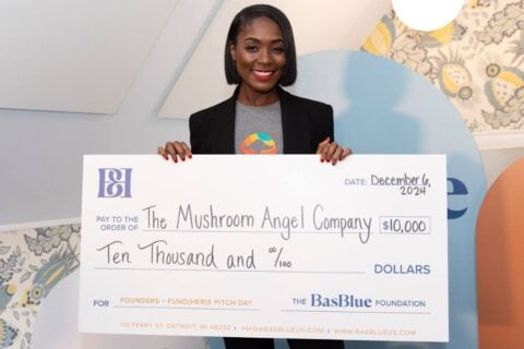A woman wearing a black blazer and a gray t-shirt with a colorful mushroom graphic smiles as she holds a large novelty check. The check is made out to “The Mushroom Angel Company” for $10,000 and is dated December 6, 2024. The check is from The BasBlue Foundation and is labeled for “Founders + Fund(HER)s Pitch Day.”