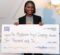 A woman wearing a black blazer and a gray t-shirt with a colorful mushroom graphic smiles as she holds a large novelty check. The check is made out to “The Mushroom Angel Company” for $10,000 and is dated December 6, 2024. The check is from The BasBlue Foundation and is labeled for “Founders + Fund(HER)s Pitch Day.”