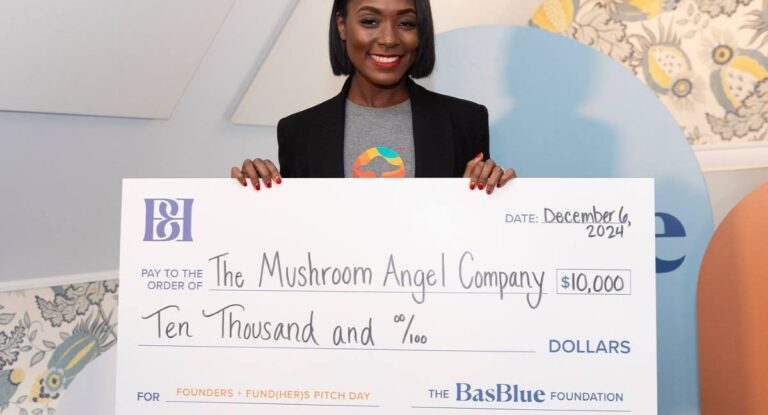 A woman wearing a black blazer and a gray t-shirt with a colorful mushroom graphic smiles as she holds a large novelty check. The check is made out to “The Mushroom Angel Company” for $10,000 and is dated December 6, 2024. The check is from The BasBlue Foundation and is labeled for “Founders + Fund(HER)s Pitch Day.”