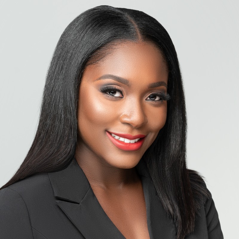 Professional headshot of a woman with long, straight black hair who is wearing a black blazer and bright red lipstick. She is smiling warmly against a plain light-gray background.
