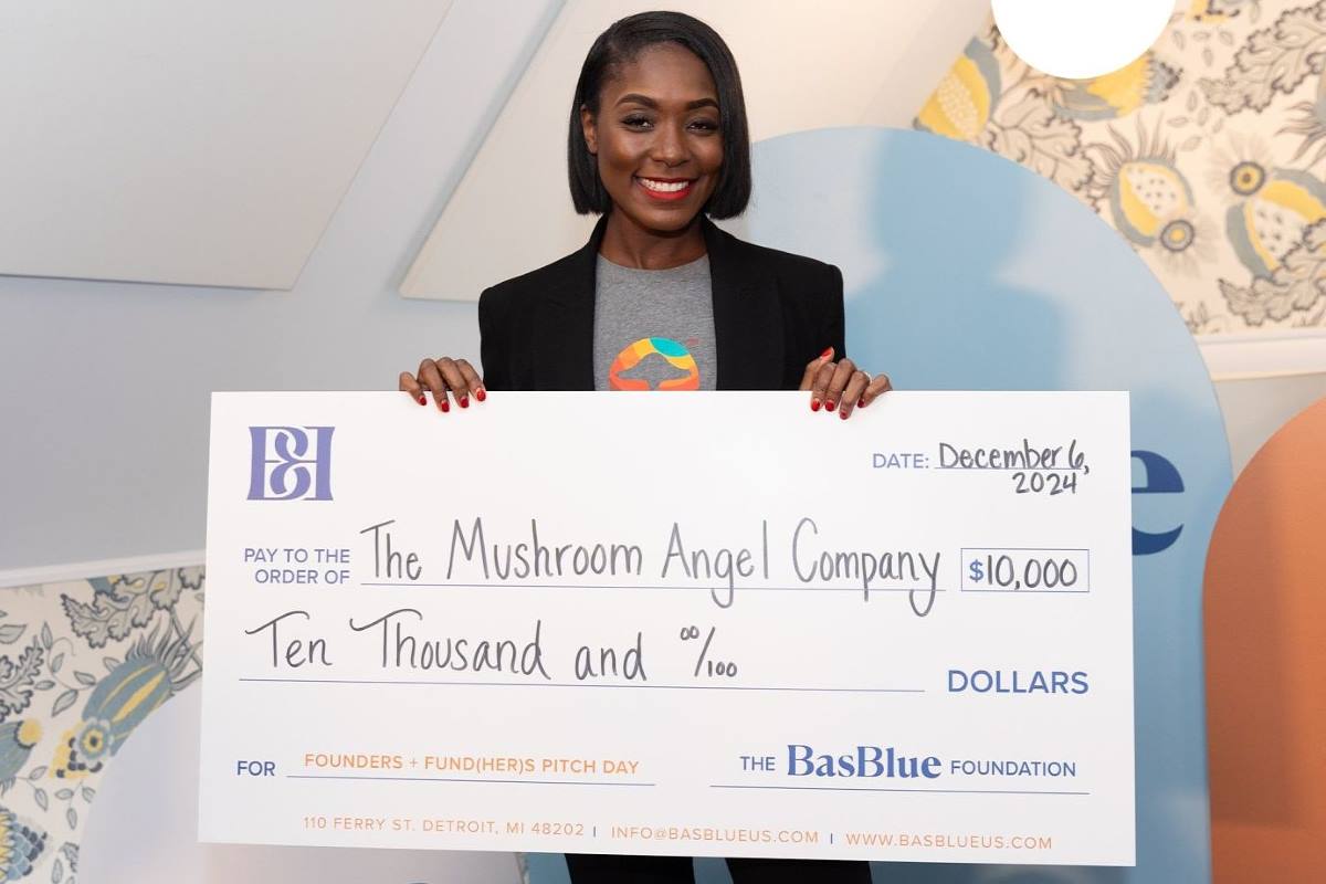 A woman wearing a black blazer and a gray t-shirt with a colorful mushroom graphic smiles as she holds a large novelty check. The check is made out to “The Mushroom Angel Company” for $10,000 and is dated December 6, 2024. The check is from The BasBlue Foundation and is labeled for “Founders + Fund(HER)s Pitch Day.”