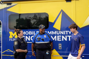Two uniformed UM-Flint DPS police officers and a member of UM-Flint's Esports team standing in front of the community engagement van, which is vinyl wrapped in maize and blue.