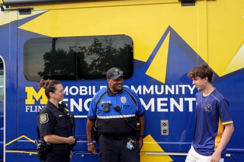 Two uniformed UM-Flint DPS police officers and a member of UM-Flint's Esports team standing in front of the community engagement van, which is vinyl wrapped in maize and blue.