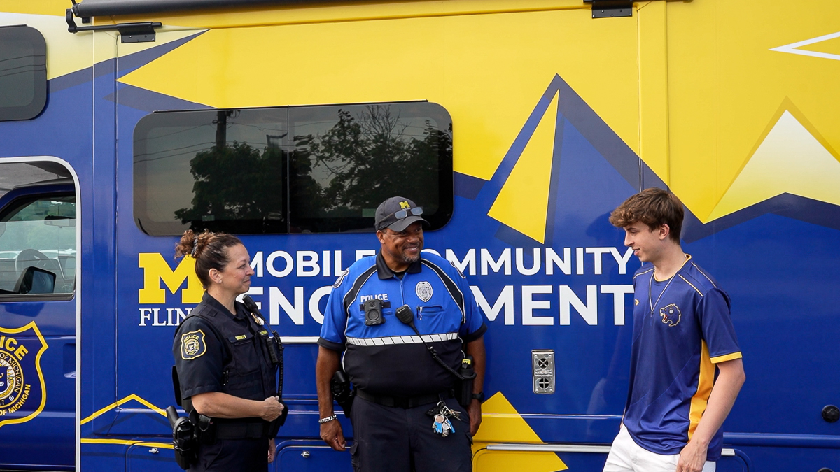 Two uniformed UM-Flint DPS police officers and a member of UM-Flint's Esports team standing in front of the community engagement van, which is vinyl wrapped in maize and blue.