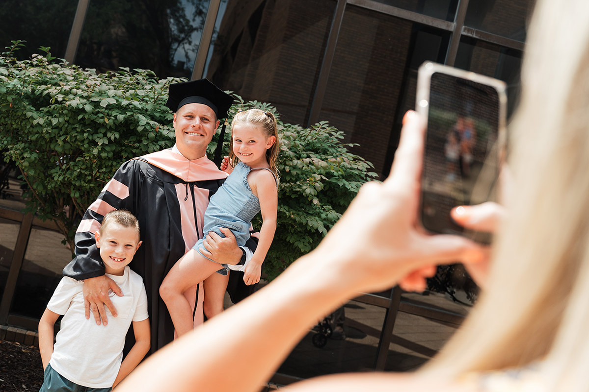 A male DNAP graduate poses for a photo while holding his kids.