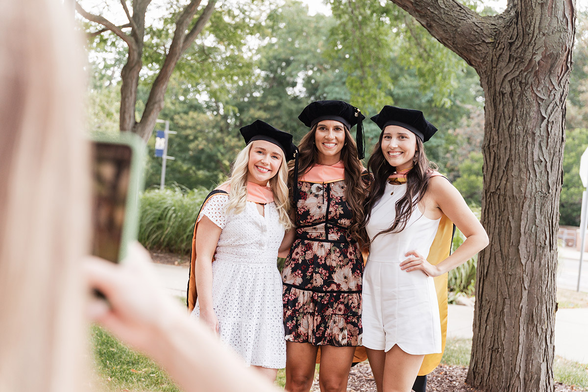 Three female DNAP graduates posing for a photo outside of the UM-Flint Theatre. They are wearing their graduation caps.