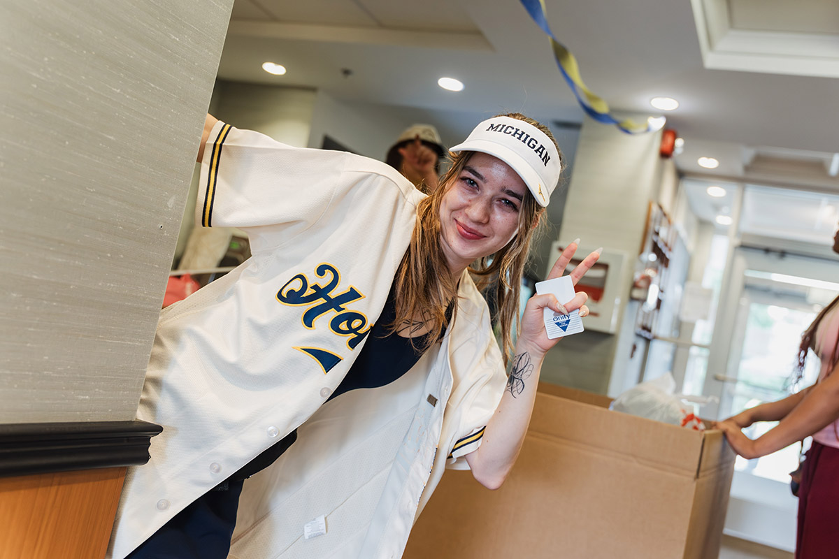 A UM-Flint student volunteer in a Michigan baseball jersey peaking from around a corner and giving the peace sign