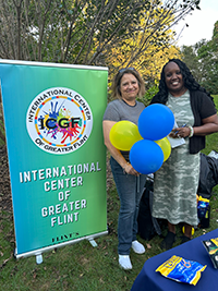 Elsa Butterfield and RoShawndra Opara stand next to a sign for the International Center of Greater Flint at a park during a community event