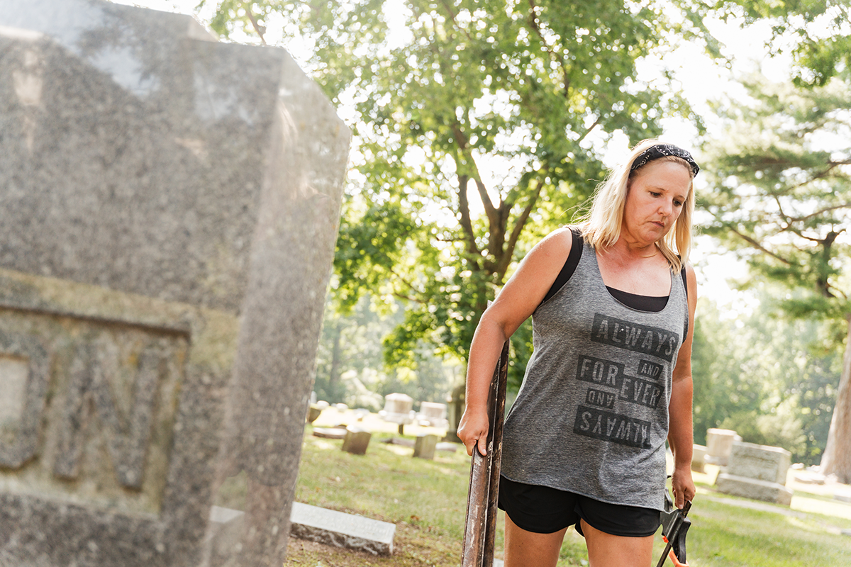 Melanie Purkis walking by a gravestone at Glenwood Cemetery wearing a gray tank top and black shorts