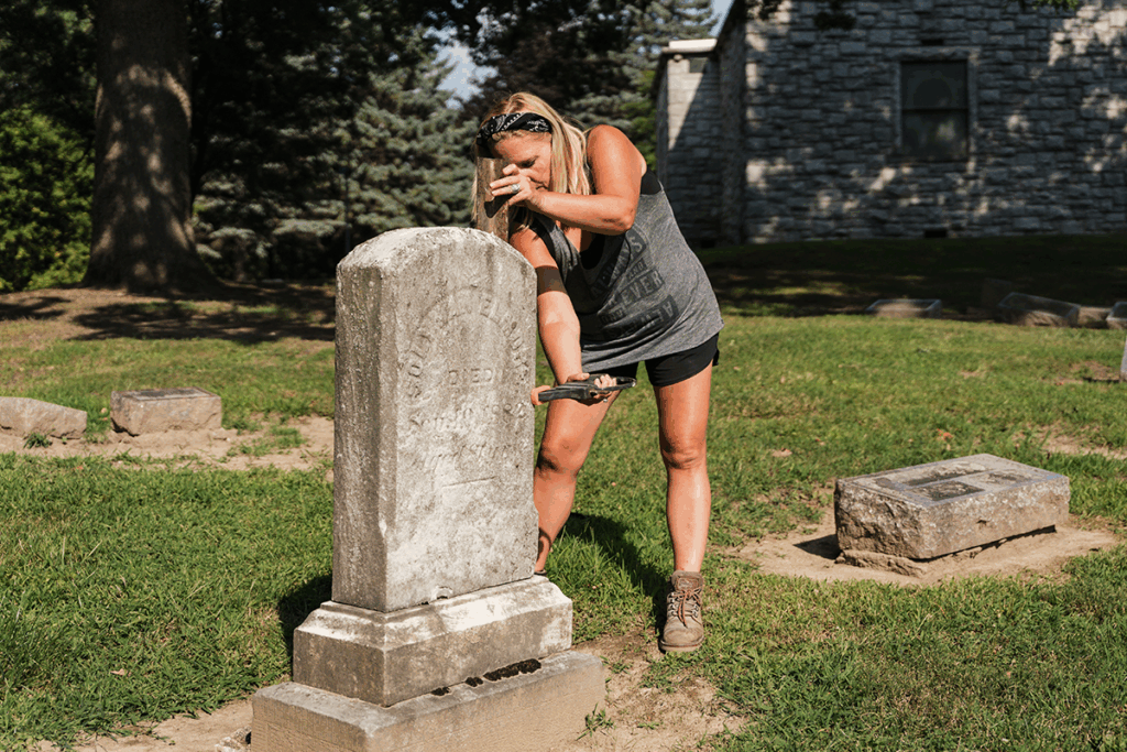 Melanie Purkis stands over a gravestone that she is repairing. She holds tools that enable her to straighten and adjust the stone face. It is a summer day at Glenwood Cemetery.