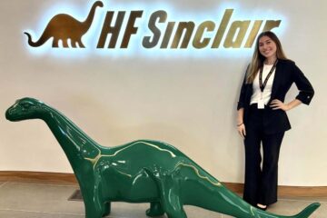 A young woman wearing a black suit with a white shirt and a lanyard around her neck stands confidently with one hand on her hip next to DINO, a shiny, dark-green dinosaur statue. Behind them, a glowing wall sign reads “HF Sinclair” with a brown dinosaur logo to the left of the text. The setting is the company's Dallas, Texas, headquarters, which features neutral-toned tile flooring and a minimalist design.