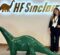 A young woman wearing a black suit with a white shirt and a lanyard around her neck stands confidently with one hand on her hip next to DINO, a shiny, dark-green dinosaur statue. Behind them, a glowing wall sign reads “HF Sinclair” with a brown dinosaur logo to the left of the text. The setting is the company's Dallas, Texas, headquarters, which features neutral-toned tile flooring and a minimalist design.