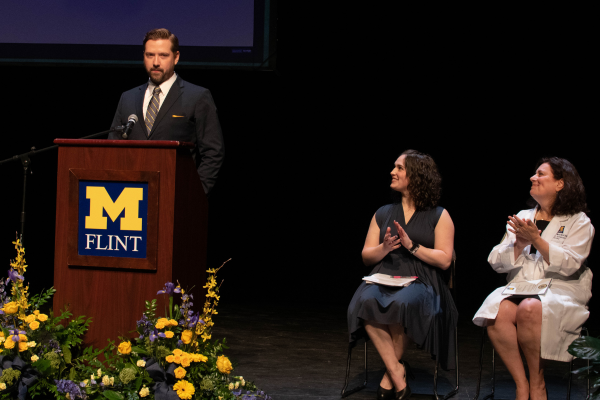 Keith Saylor, in a suit, speaks at a lectern on stage. Two UM-Flint faculty members are in view seated on stage. They are clapping and looking at Keith.