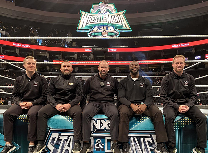 Clayton Tomczyk (far left) and his fellow WWE PT therapists sit on the side of a wrestling ring, smiling.