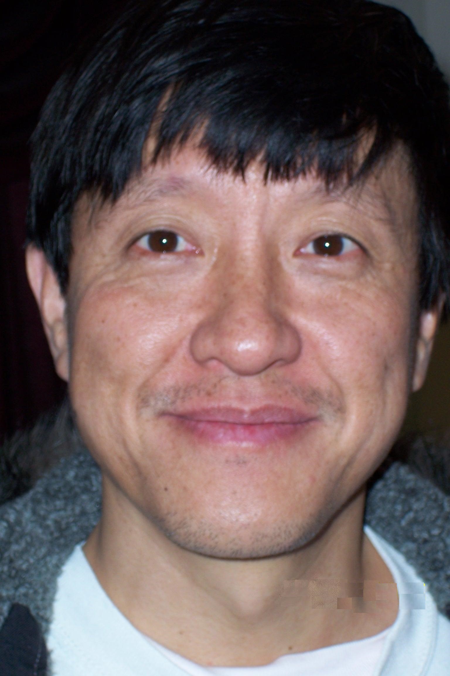A close-up portrait of a man with straight, dark hair and a short, thin mustache. He is smiling slightly, looking directly at the camera. He is wearing a white shirt under a grey jacket, and the background is dark, keeping the focus on his face.