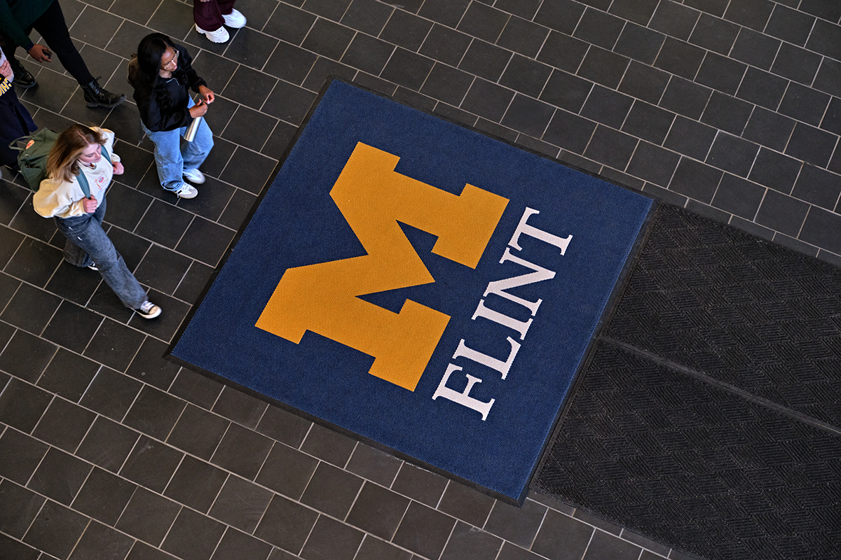 An overhead view of the first floor of the University Center with students walking close to a UM-Flint stamp logo on the floor.