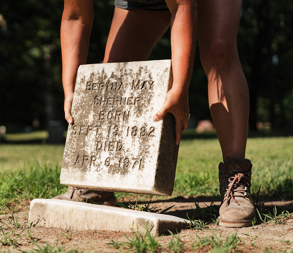 A close up of Purkis holding a headstone close to the ground. The headstone reads Bertha May Shepner. Born Sept. 13 1882. Died Apr 6 1971