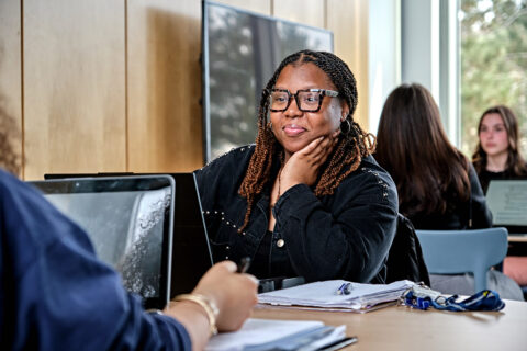 A female student sitting at a desk talking to another student who is mostly off camera. Two more students are in the background. Also in the background is a window looking out to a sunny day with trees.