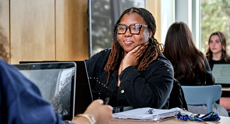 A female student sitting at a desk talking to another student who is mostly off camera. Two more students are in the background. Also in the background is a window looking out to a sunny day with trees.