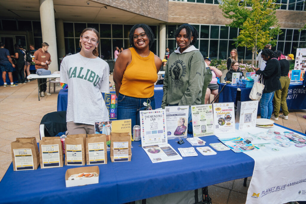 Three women stand behind the Planet Blue Ambassador table to promote sustainability on campus.