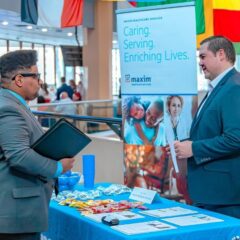 A young man in a suit speaks with a recruiter at a Maxim Healthcare Services booth during a Career Fair. The booth has a blue tablecloth with candy, flyers and giveaways. A banner behind the recruiter reads “Caring. Serving. Enriching Lives.” atop photos of healthcare professionals and families. International flags hang in the background near tall windows.