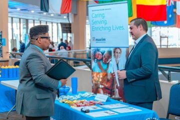 A young man in a suit speaks with a recruiter at a Maxim Healthcare Services booth during a Career Fair. The booth has a blue tablecloth with candy, flyers and giveaways. A banner behind the recruiter reads “Caring. Serving. Enriching Lives.” atop photos of healthcare professionals and families. International flags hang in the background near tall windows.