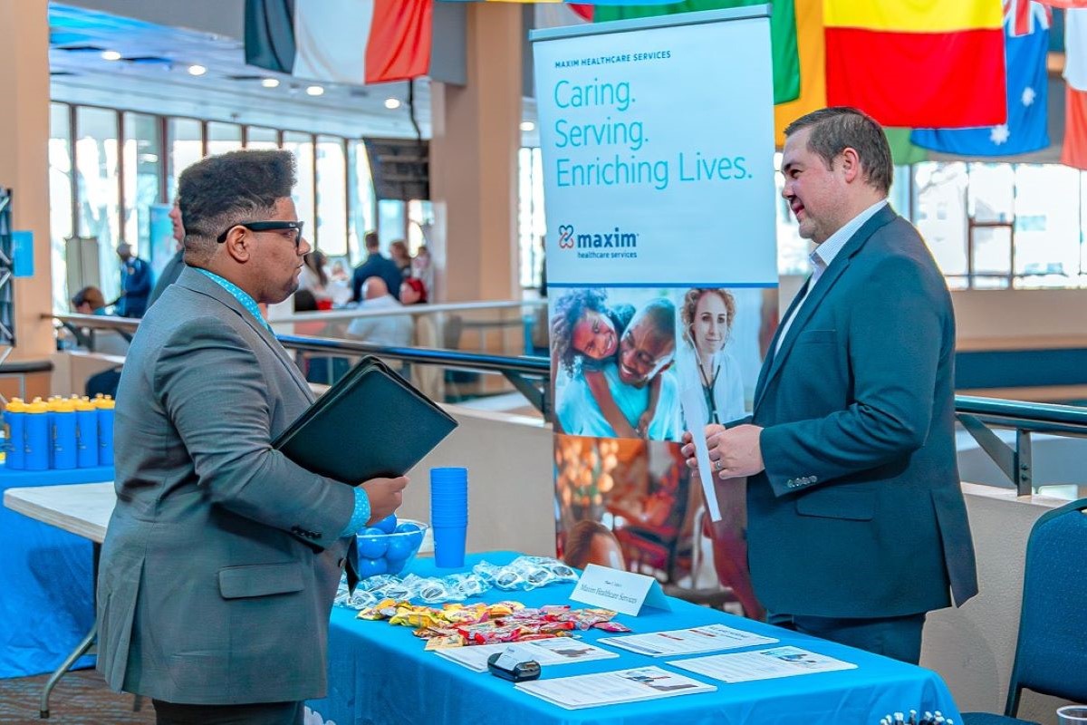 A young man in a suit speaks with a recruiter at a Maxim Healthcare Services booth during a Career Fair. The booth has a blue tablecloth with candy, flyers and giveaways. A banner behind the recruiter reads “Caring. Serving. Enriching Lives.” atop photos of healthcare professionals and families. International flags hang in the background near tall windows.