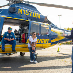 The University of Michigan Survival Flight Helicopter in a parkimg lot. A member of the flight crew is sitting in helicopter, while a mom and son pose for a photo.