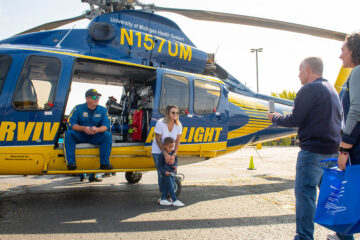 The University of Michigan Survival Flight Helicopter in a parkimg lot. A member of the flight crew is sitting in helicopter, while a mom and son pose for a photo.