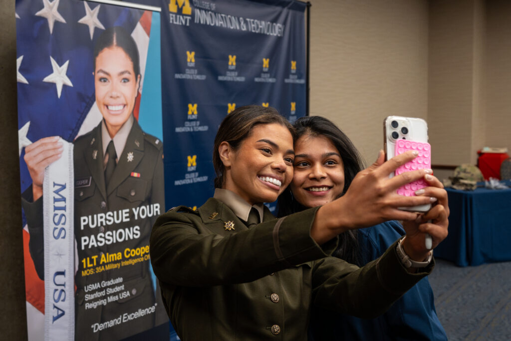 Alma Cooper in military uniform holding a cell phone to take a photo, smiling with a female student. 