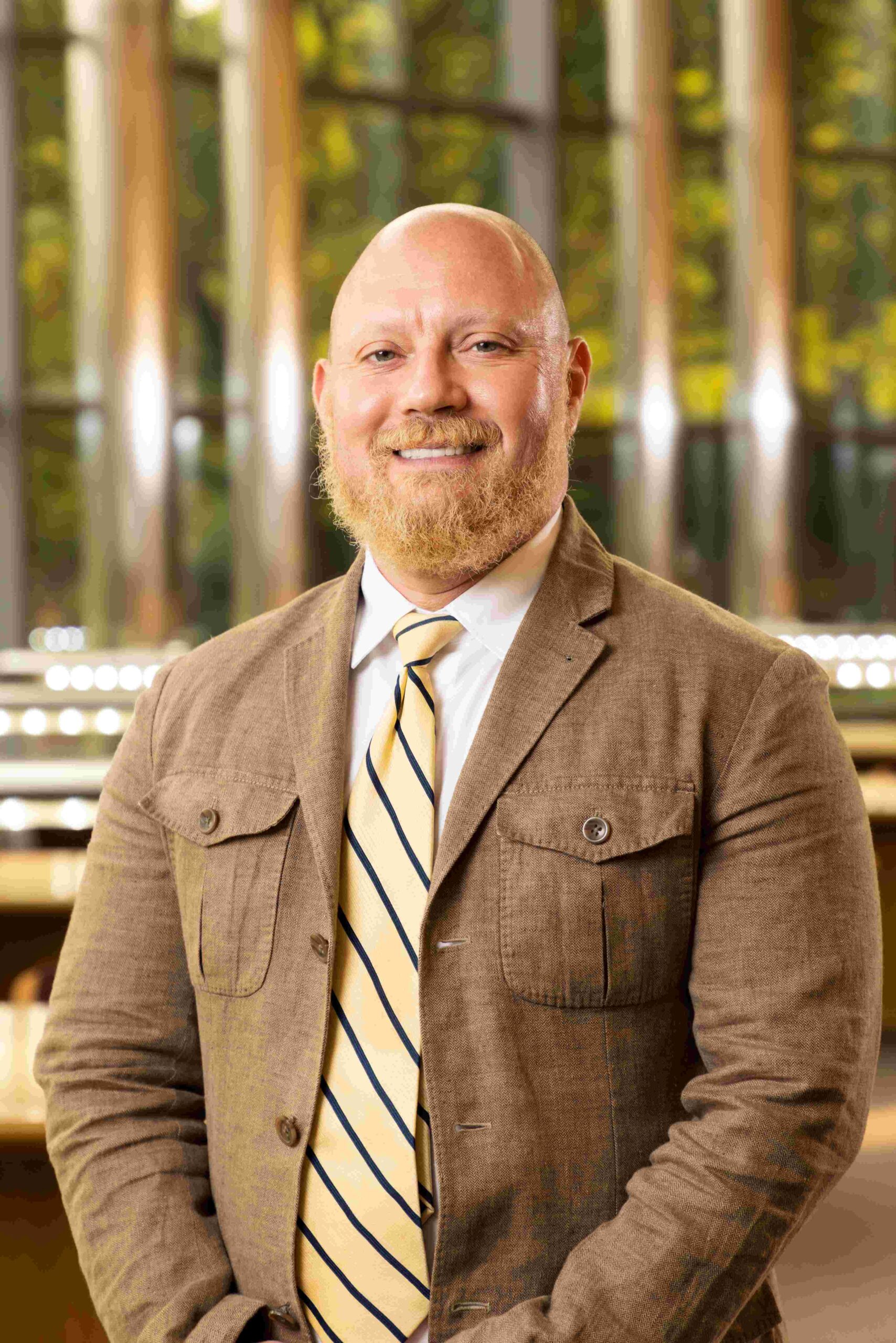 A bald man with a blond beard smiles warmly. He is wearing a brown jacket, white dress shirt, and yellow tie with diagonal, navy-blue stripes. The background features tall glass windows with green foliage visible outside and bright interior lighting.
