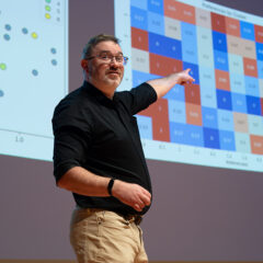 Cam McLeman speaking on stage, pointing to graphs projected on the wall behind him