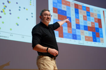 Cam McLeman speaking on stage, pointing to graphs projected on the wall behind him