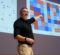 Cam McLeman speaking on stage, pointing to graphs projected on the wall behind him