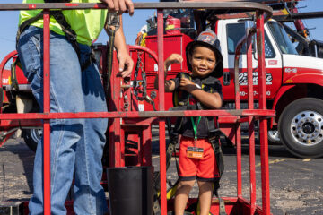 A young boy is flexing his muscles while wearing a fireman's helmet and standing in a cherry picker bucket