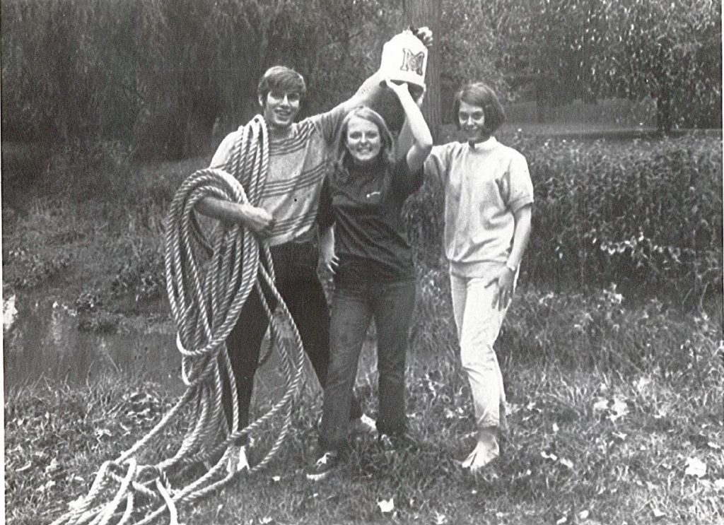 Alumni David Zick (left), Tina Peters (middle), and Barb Tomcal (right), in a black and white photo holding a rope and trophy. 