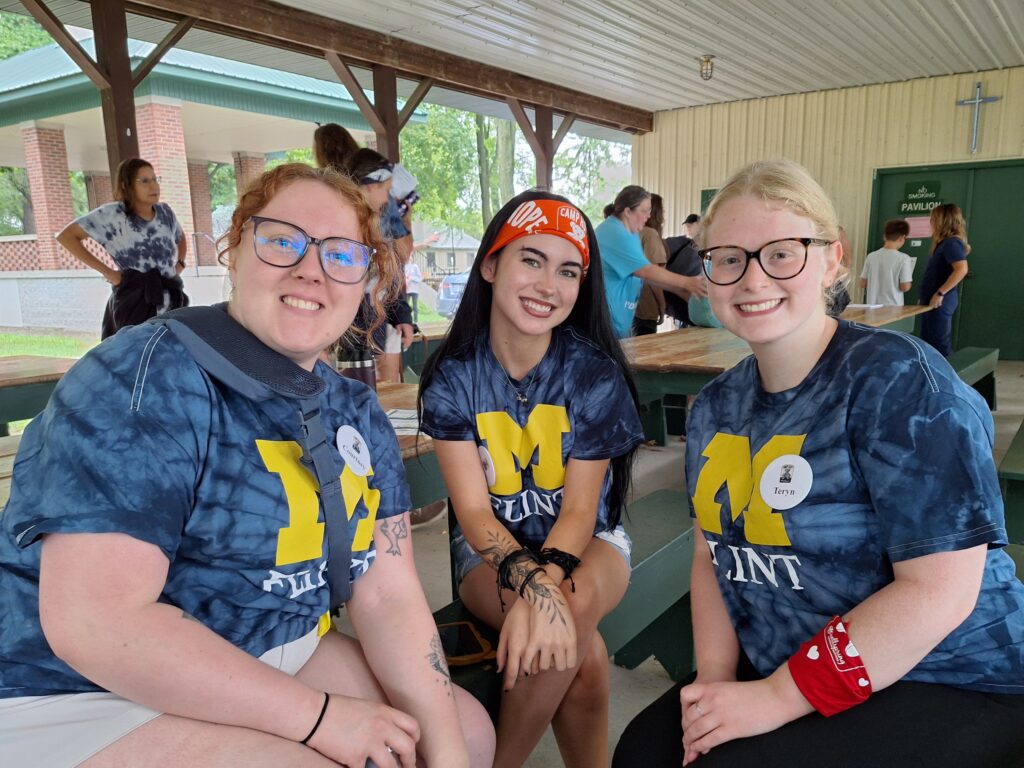 Three young women sit in an outdoor shelter smiling at the camera, wearing UM-Flint t-shirts