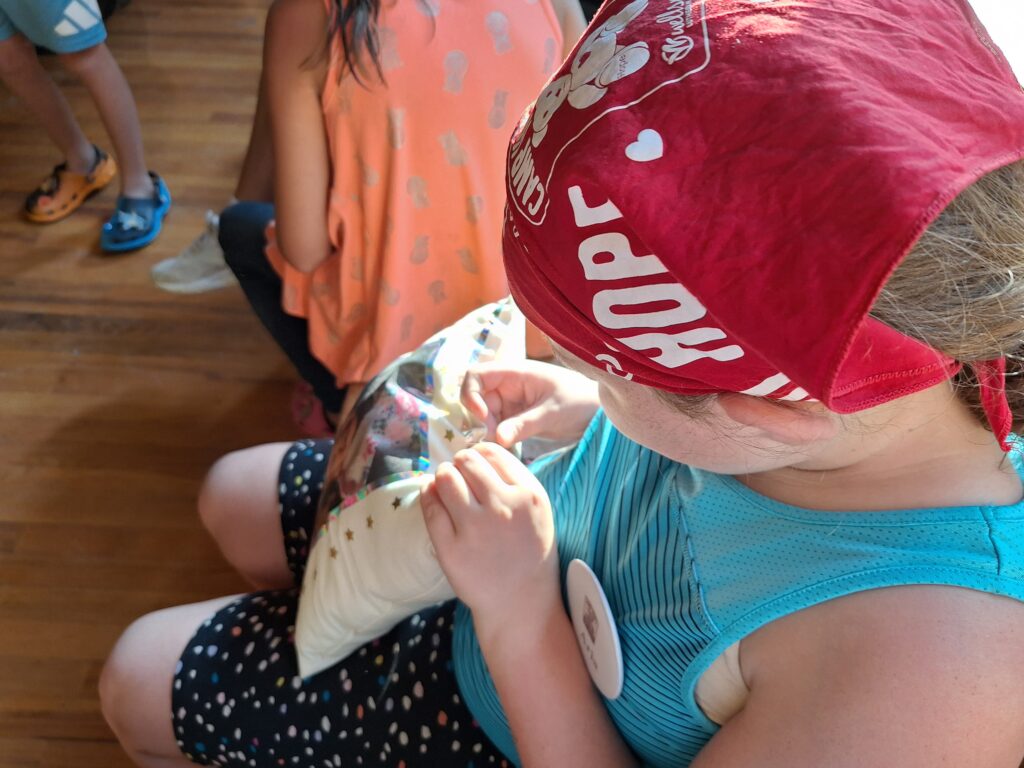 A young girl wearing a red bandana and a turquoise tank top is sewing a pillow as part of the camp's Memory Pillow activity
