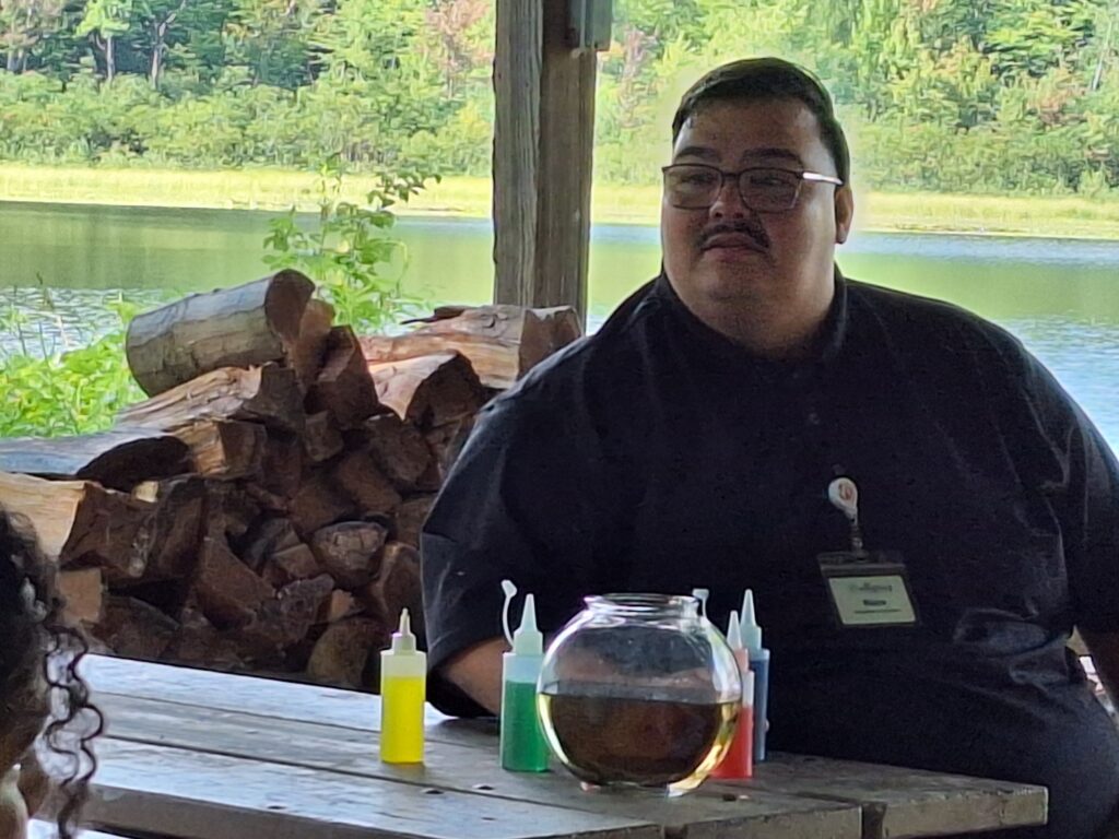 Ricco Ruiz sits at a picnic table with a log pile and lake the background