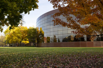The Thompson Library in Fall. A modern building with a curved glass facade surrounded by colorful autumn trees and fallen leaves on the grass.