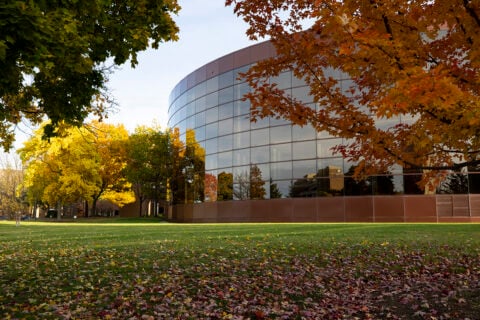 The Thompson Library in Fall. A modern building with a curved glass facade surrounded by colorful autumn trees and fallen leaves on the grass.