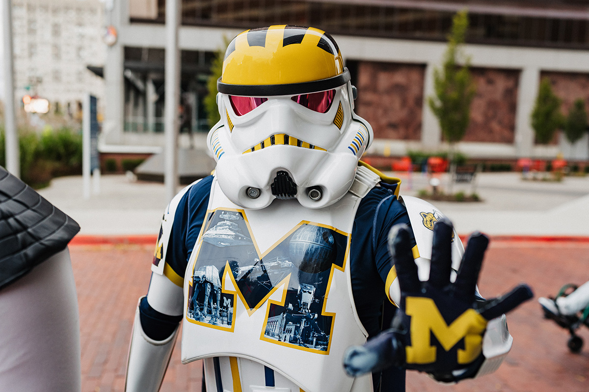 A person dressed in a stormtrooper costume with a yellow and blue design, featuring a large "M" on the chest, is holding up a hand with an "M" symbol.