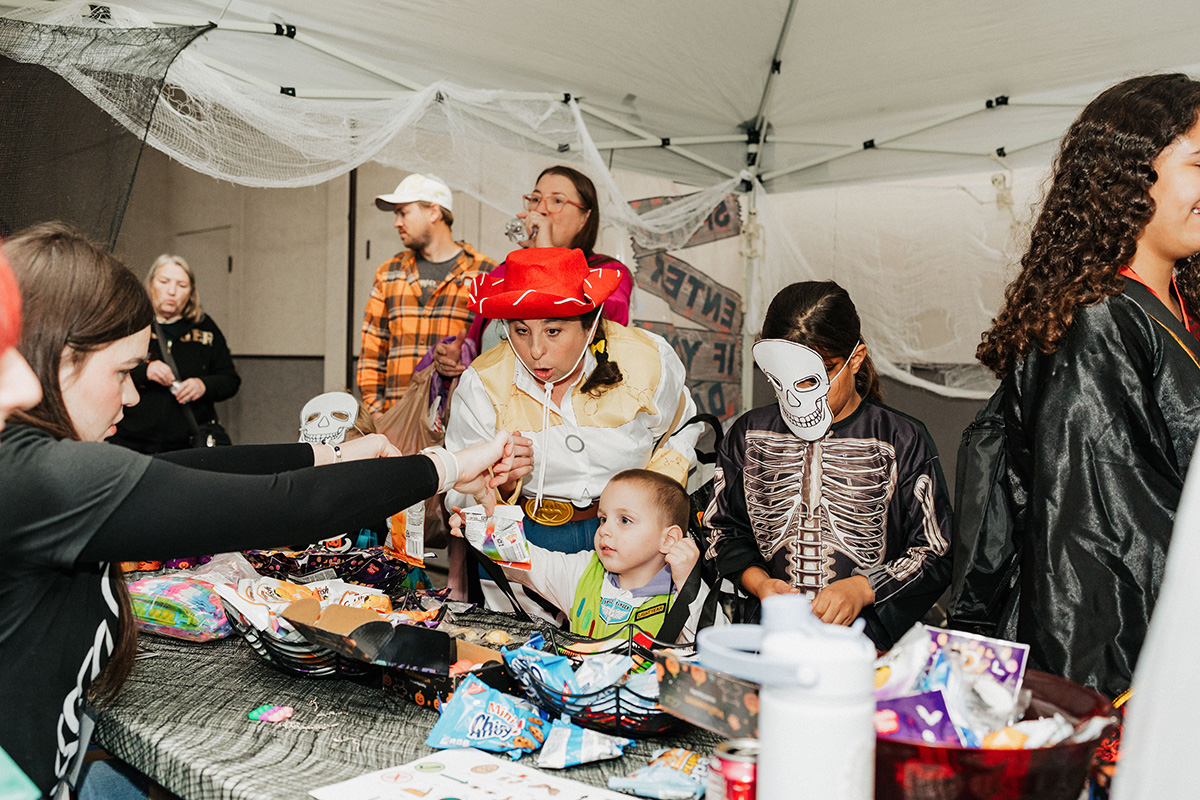 A group of children in costumes gather around a table filled with Halloween treats and decorations under a tent.