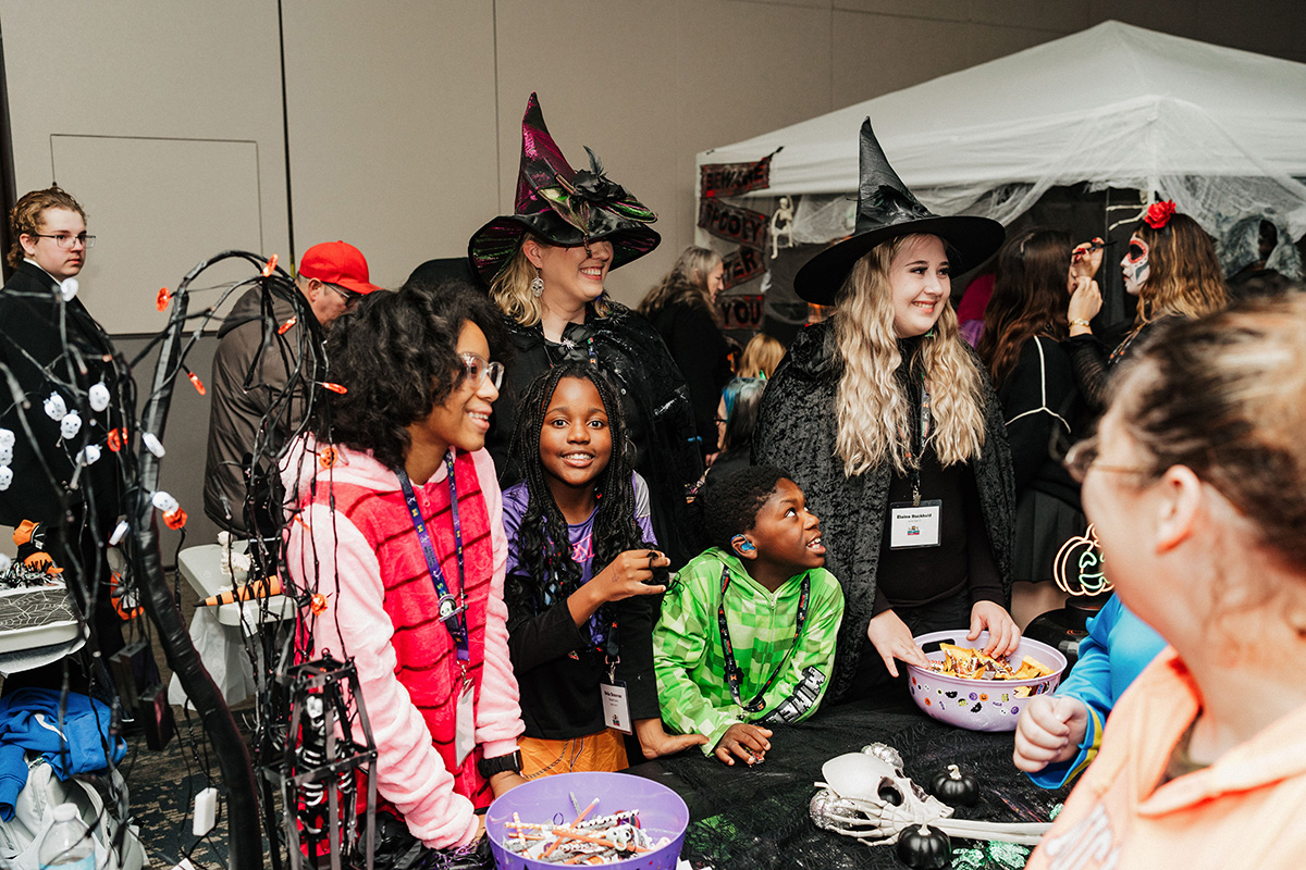 A group of children and adults in Halloween costumes gather around a table with candy and decorations.