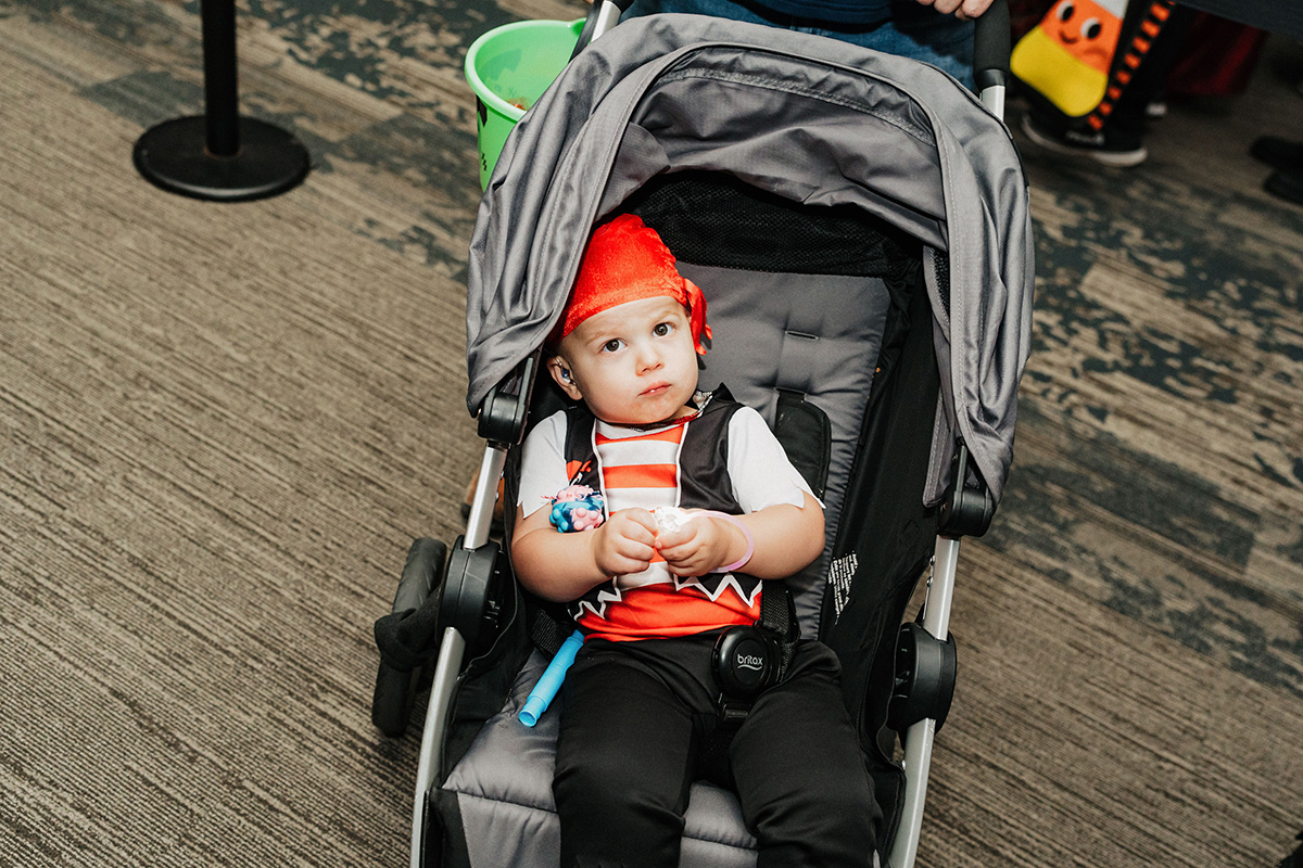 A child in a stroller wearing a costume with red and black accents, holding a toy and a blue object.