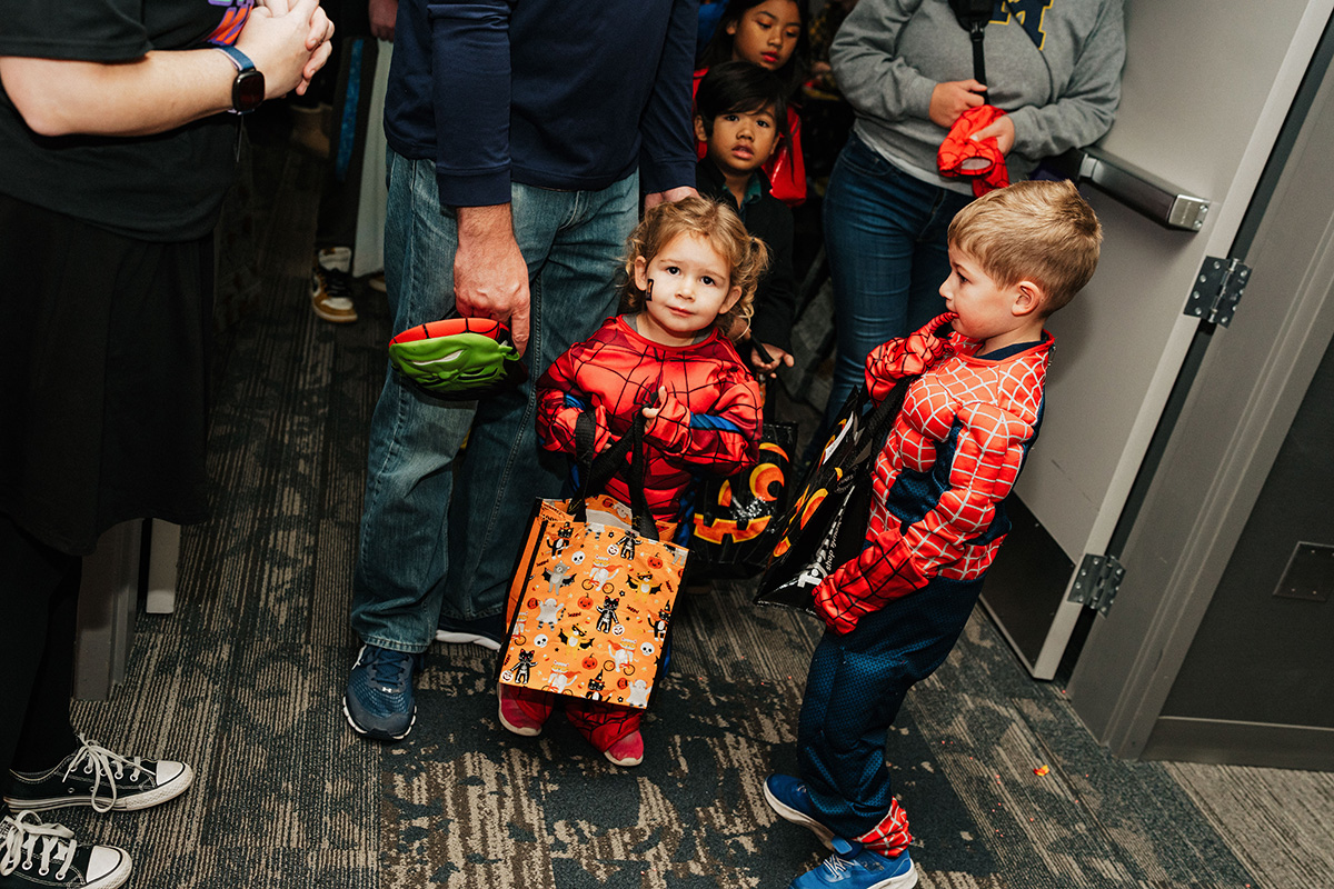 Two children dressed in Spider-Man costumes hold Halloween-themed bags while standing in a hallway.