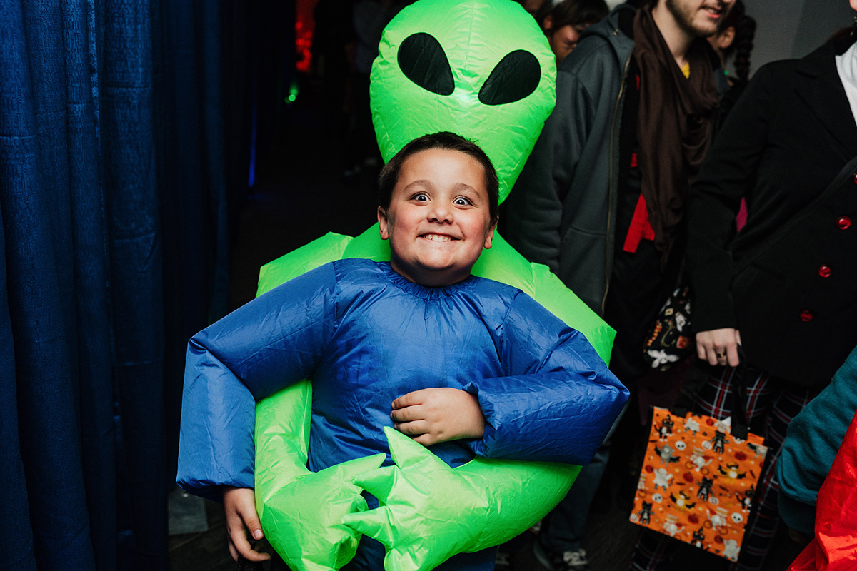 A child dressed in a blue inflatable costume being carried by a large green alien costume at a festive event.