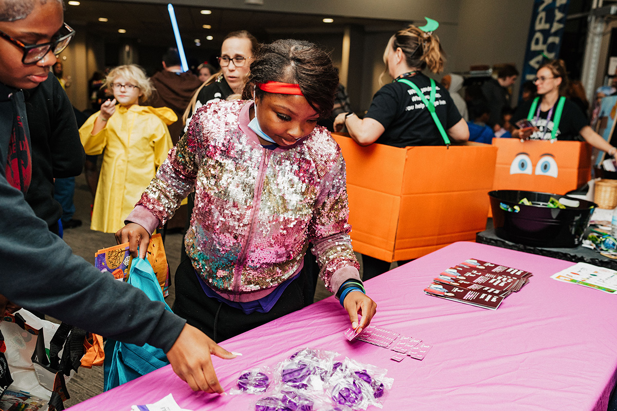 A person in a shiny, sequined jacket is reaching for purple items on a pink table at a lively event.